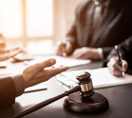 the center focus the hands of a judge, who is sitting to the left, with their gavel and block next to them, and to the right are 2 people facing the judge and writing on notepads